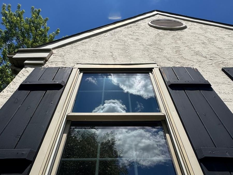 A gable window with black shutters mirroring clouds and blue sky